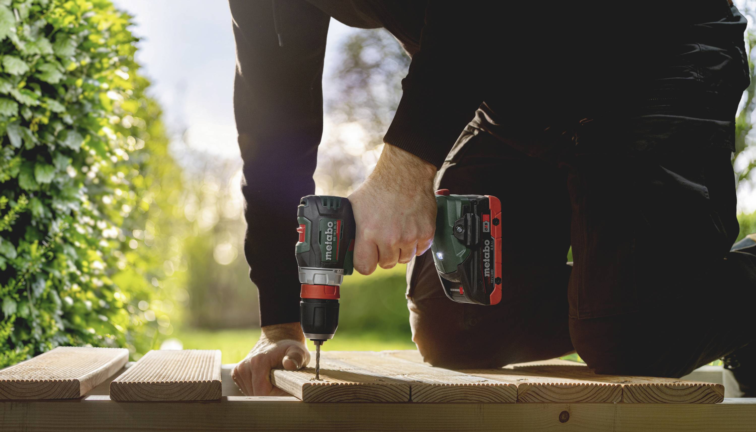 A person is kneeling outdoors and using an electric drill to screw boards together. Trees and sunshine are visible in the background.