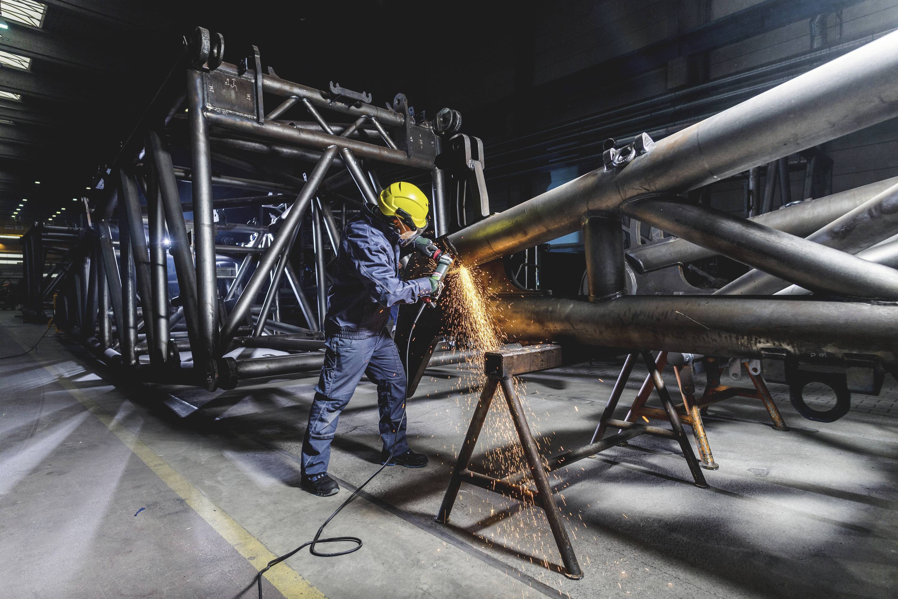 A worker wearing a yellow hard hat is grinding a metal pipe in an industrial workshop. Sparks are flying from the grinding point.
