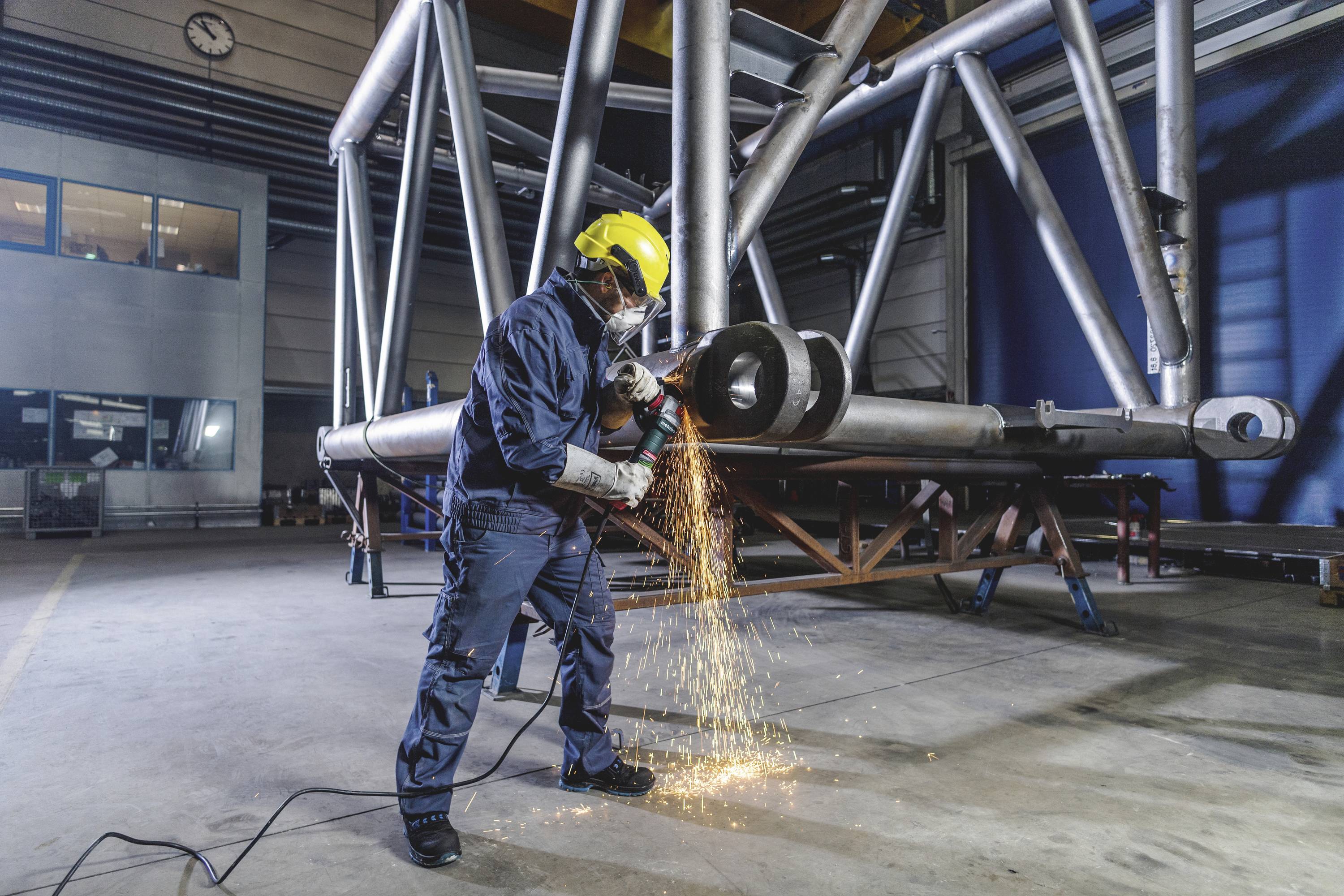A worker in protective clothing is grinding metal pipes in a factory hall. Sparks are flying as the machine processes the metal.