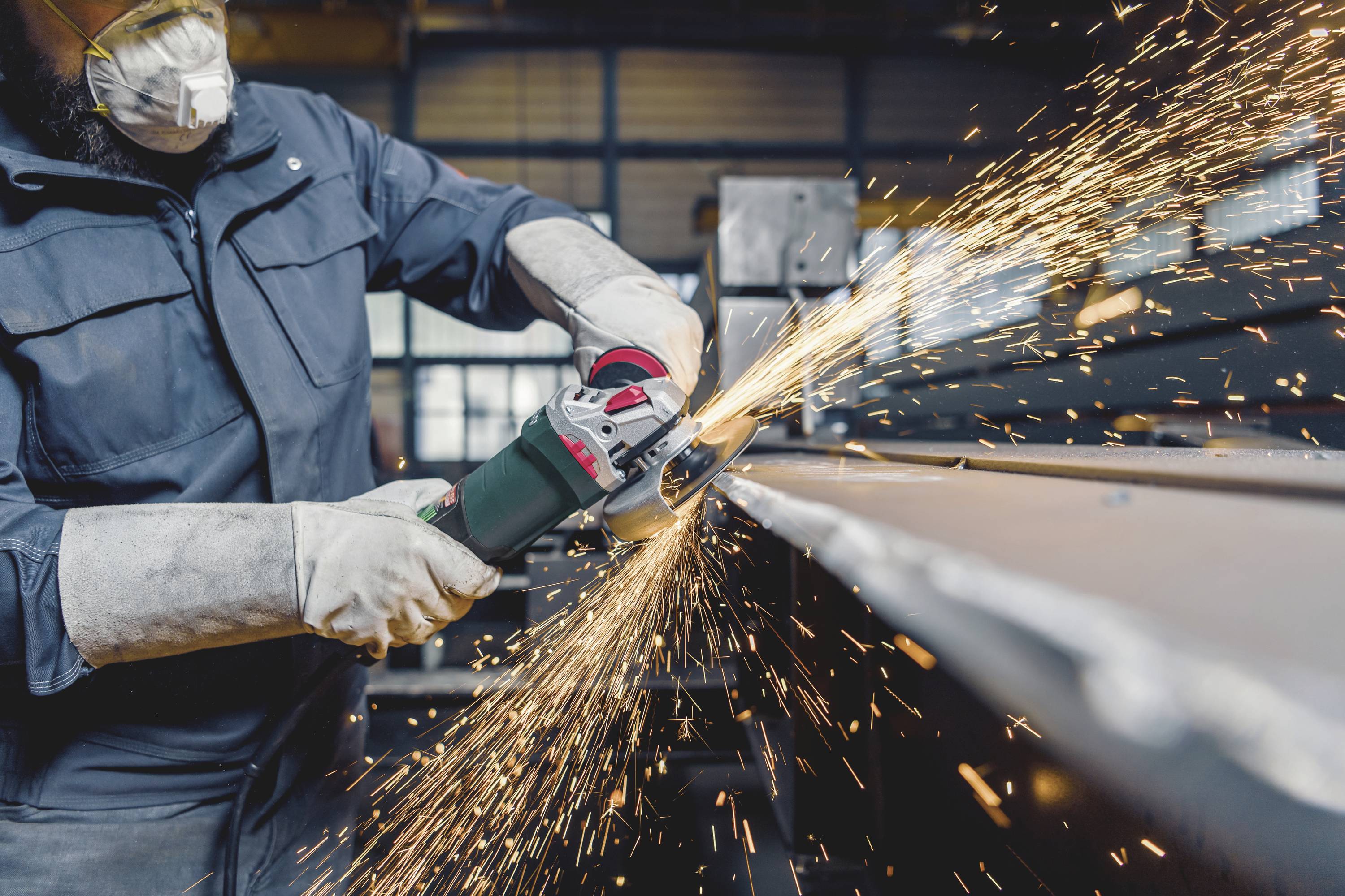 A person is grinding metal with an angle grinder, with sparks flying. Protective clothing and a mask are being worn to protect against dust and sparks.