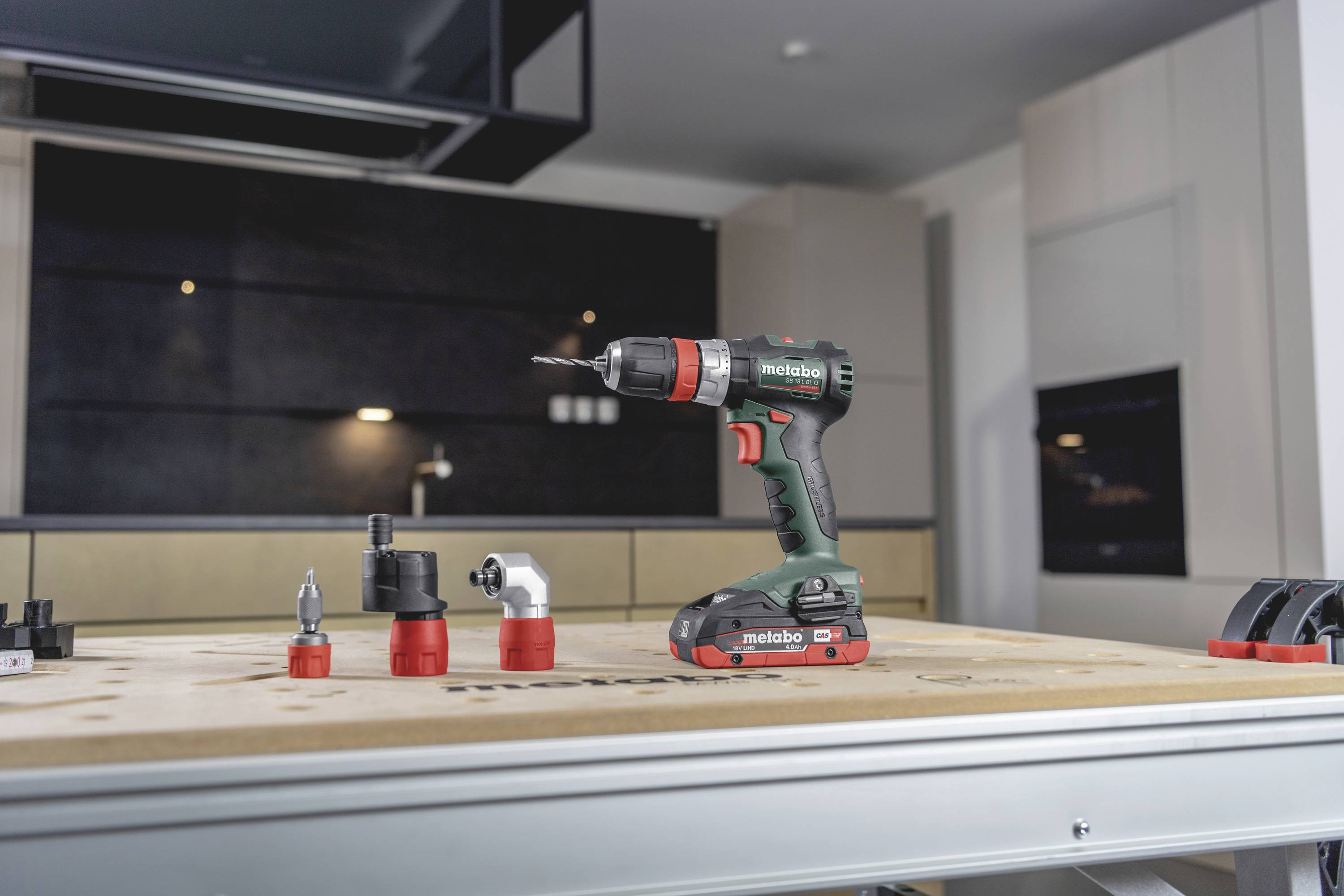 Cordless drill with various attachments on a workbench in a modern workshop. Background: dark cabinet fronts and wooden shelves.