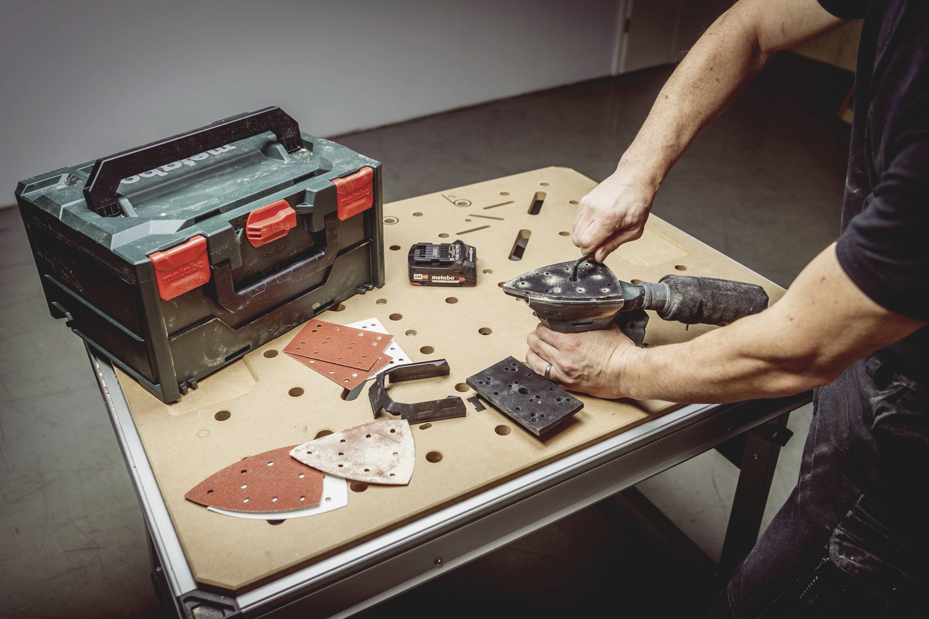A person is changing attachments on a hand-guided grinding machine on a work table. Tools are lying in a case beside it.