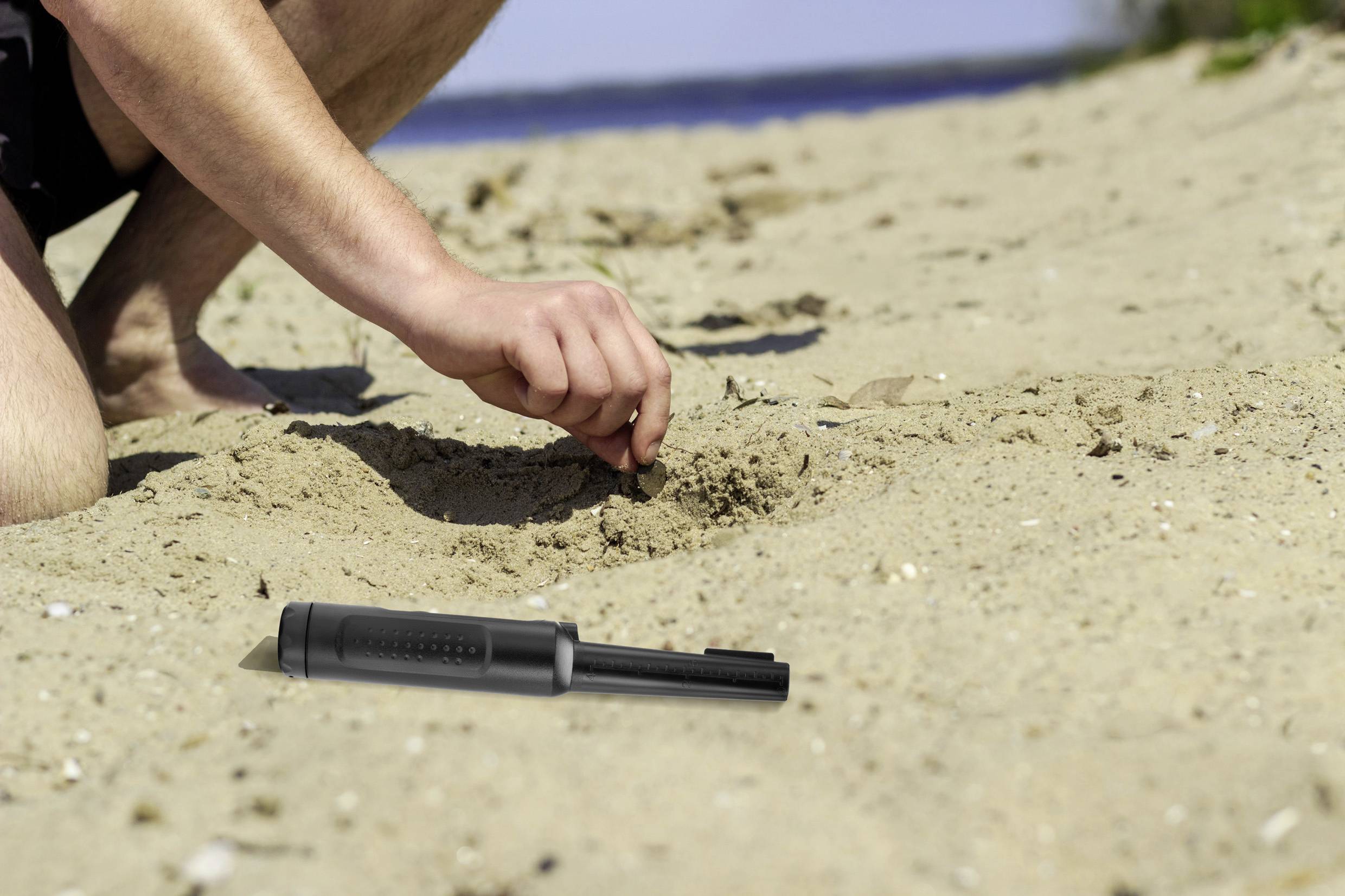 A person is digging a small hole in the sand on the beach. A black device lies beside their hand in the sand. The sea is visible in the background.