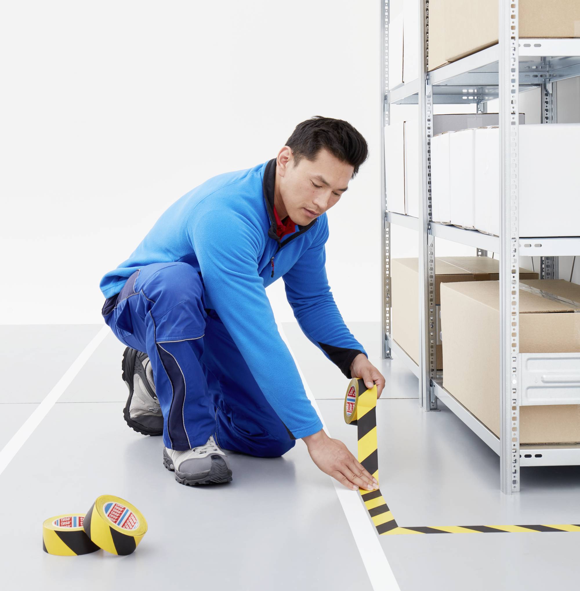 A man in blue workwear is sticking yellow and black warning tape on the floor of a warehouse next to shelves with boxes.