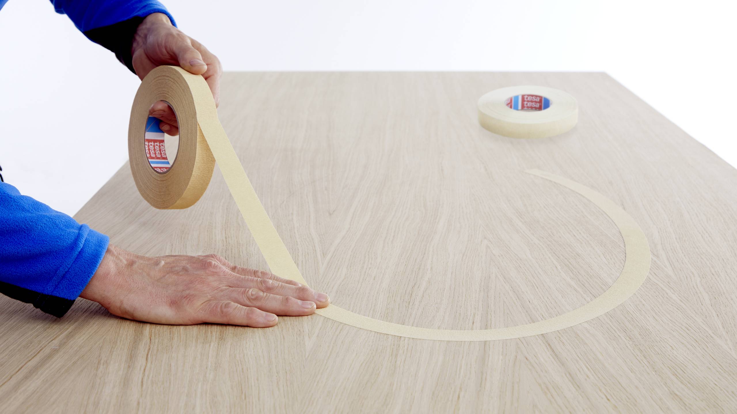 A person is sticking brown adhesive tape in a curved line onto a wooden surface. Another roll of tape lies in the background.