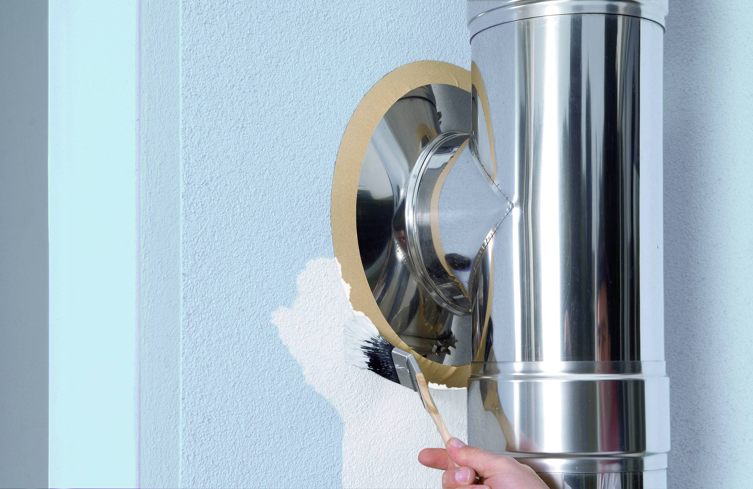 A hand is painting a wall around a shiny metal pipe, transforming the wall from white to light blue.