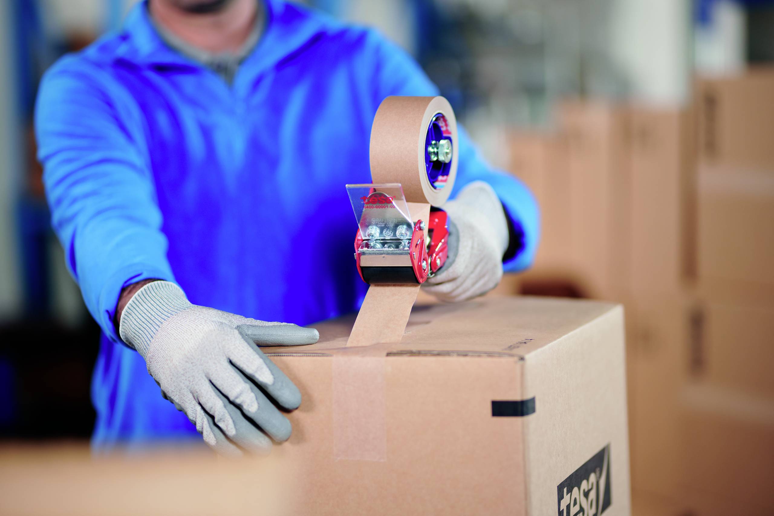 A person wearing blue clothing is sealing a cardboard box with tape in a warehouse setting. Hands are wearing gloves.