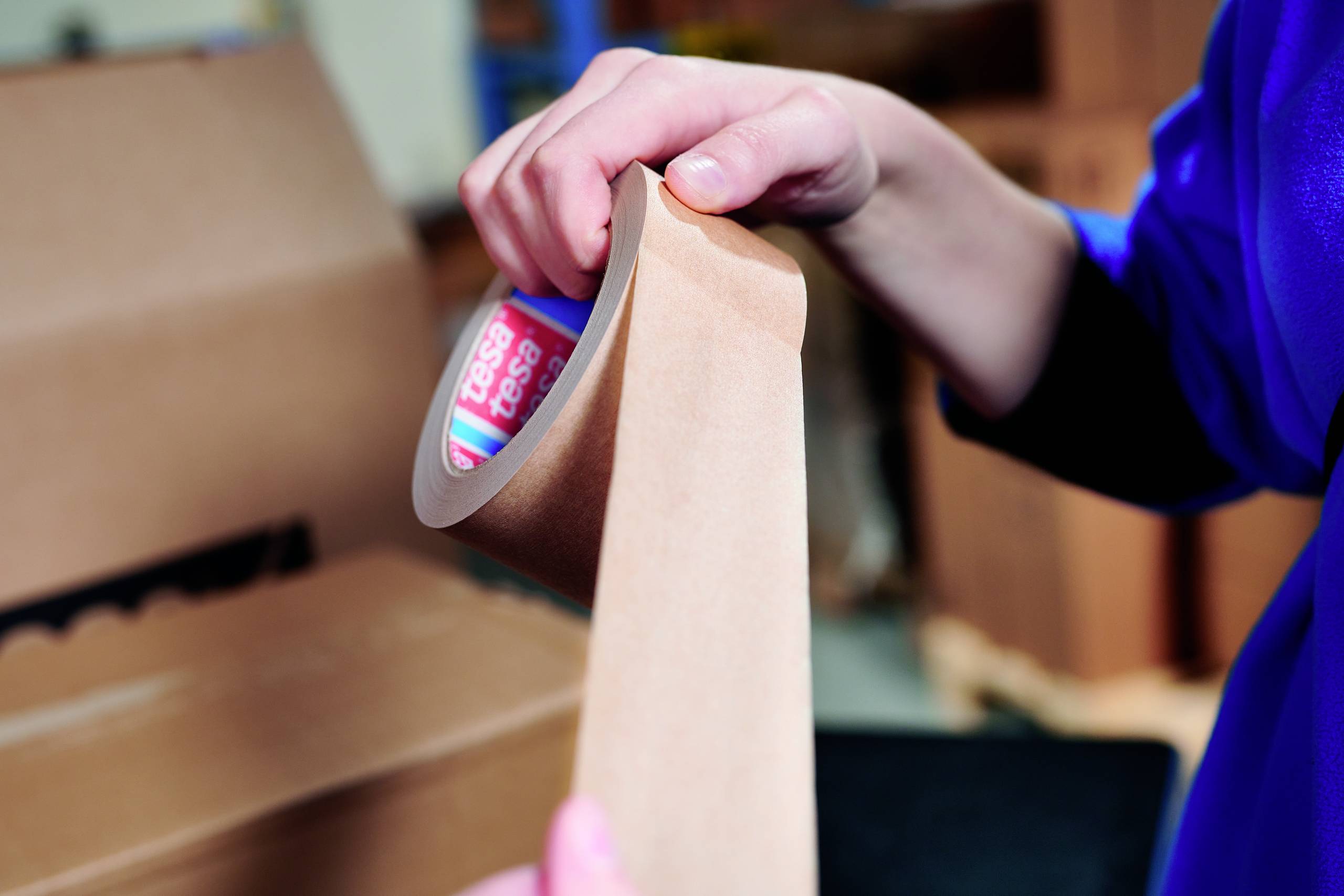 A person is holding a roll of brown packing tape and pulling a piece off it; an open cardboard box is in the background.