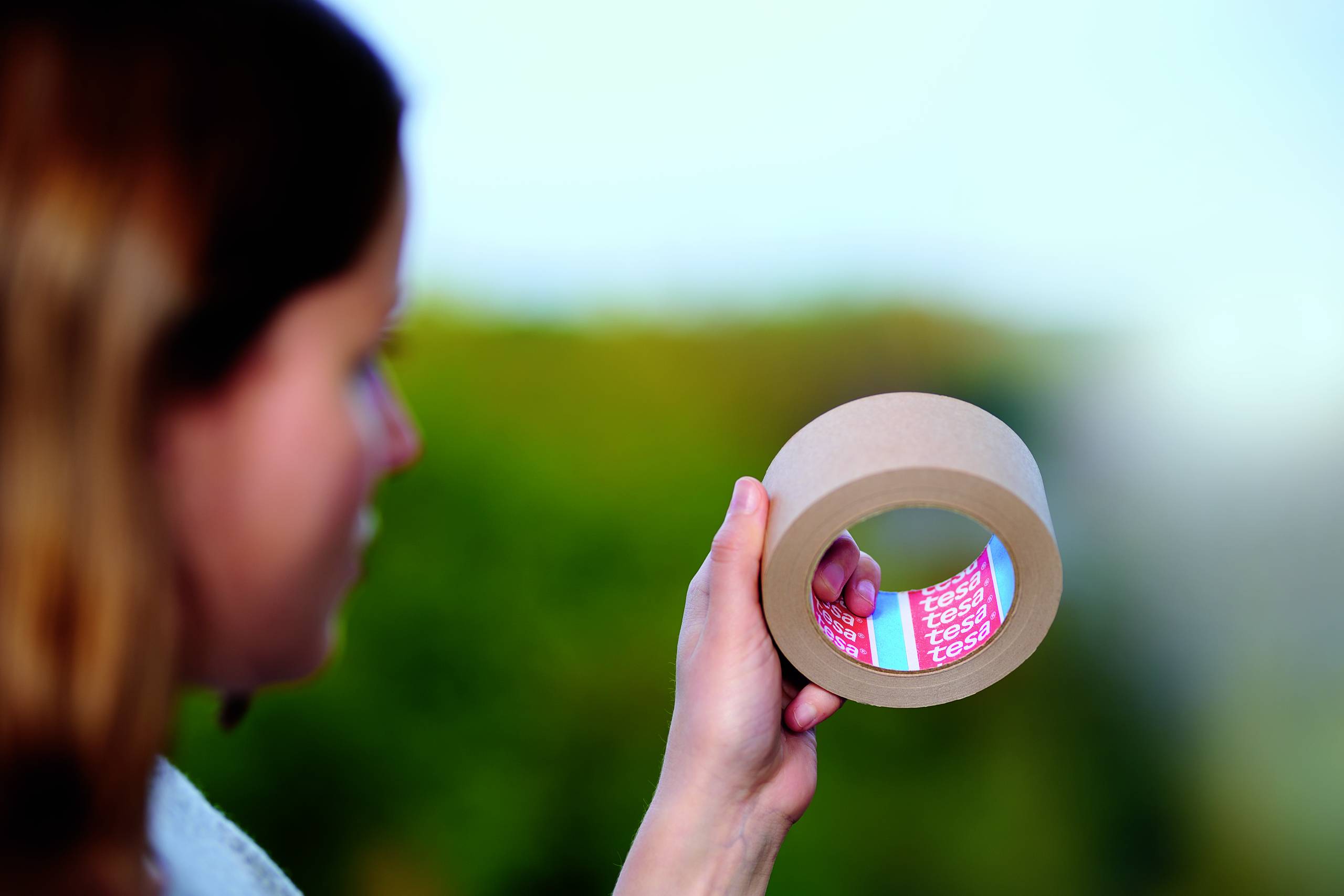 A person is holding a roll of sticky tape in their hand. The background is out of focus and shows a blurred green.