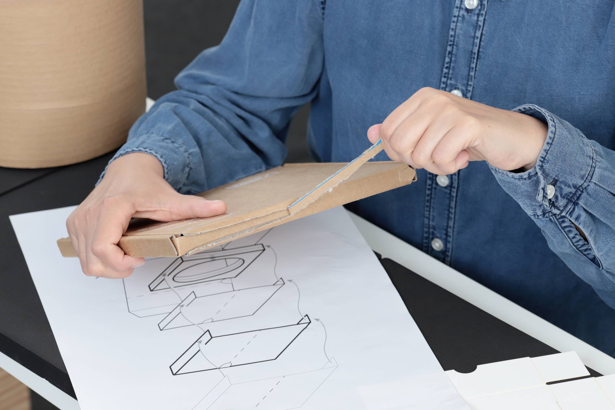 A person in a denim shirt carefully opens a package with a knife. The underlying surface shows design drawings of packaging.