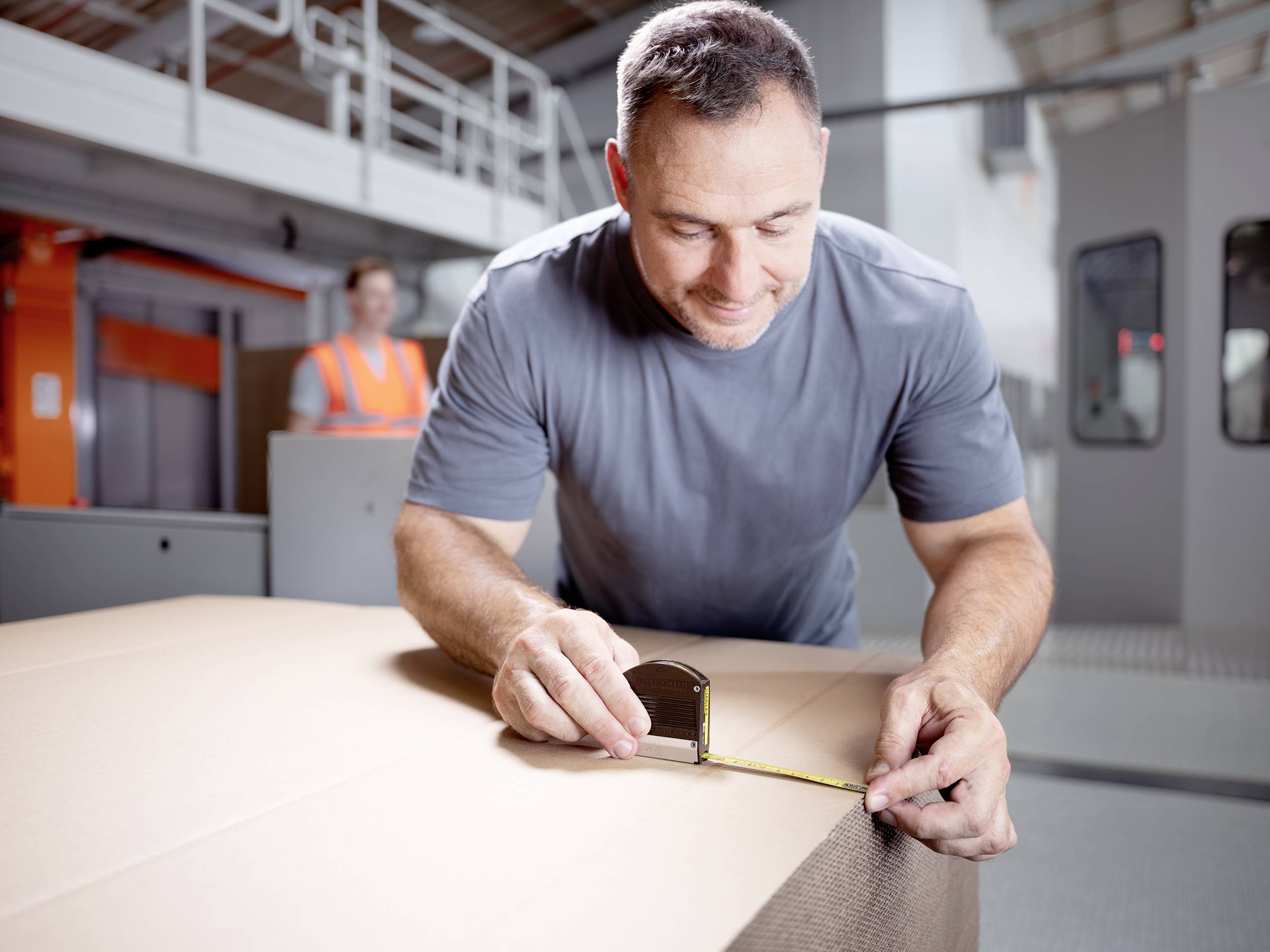 A man is measuring the edge of a large cardboard box with a tape measure in a factory setting. Another person is standing in the background.