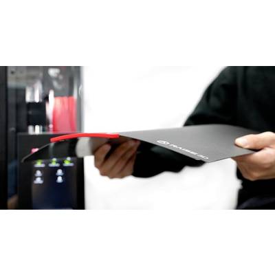 A person holds a flexible 3D printer bed in front of a 3D printer. The bed bends to easily remove a red printed object.