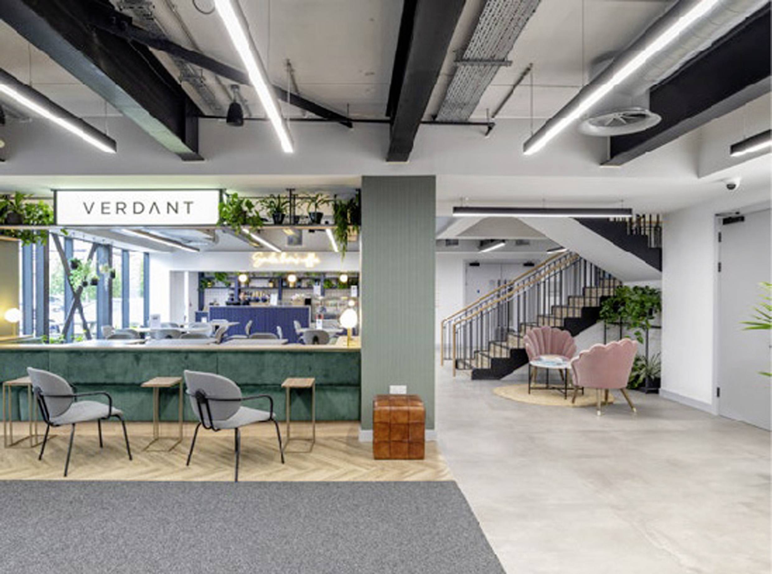 Modern office with open-plan seating areas, staircase in the background. Plants and lighting elements create an inviting atmosphere.