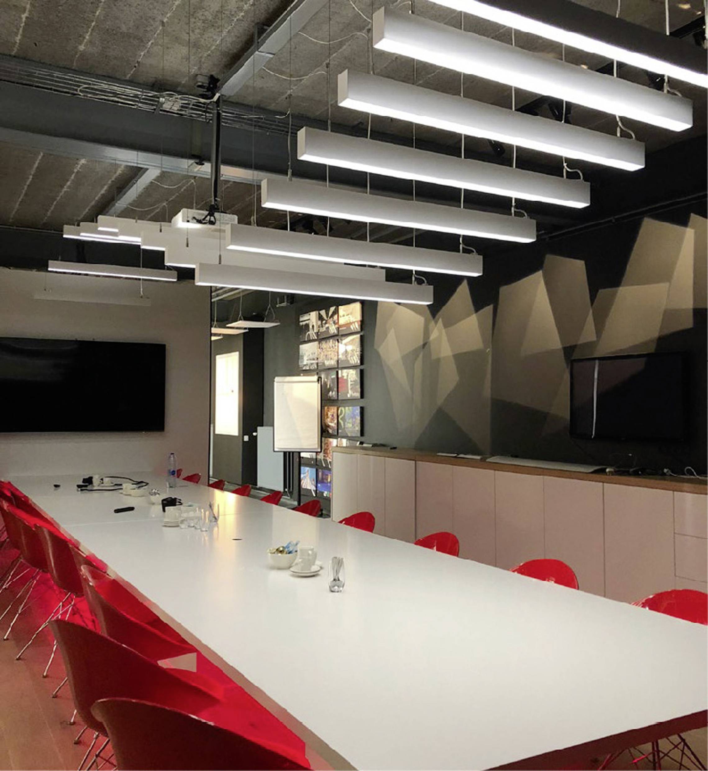 Conference room with a large white table, red chairs, pendant lights hanging from the ceiling, and a large screen on the wall.