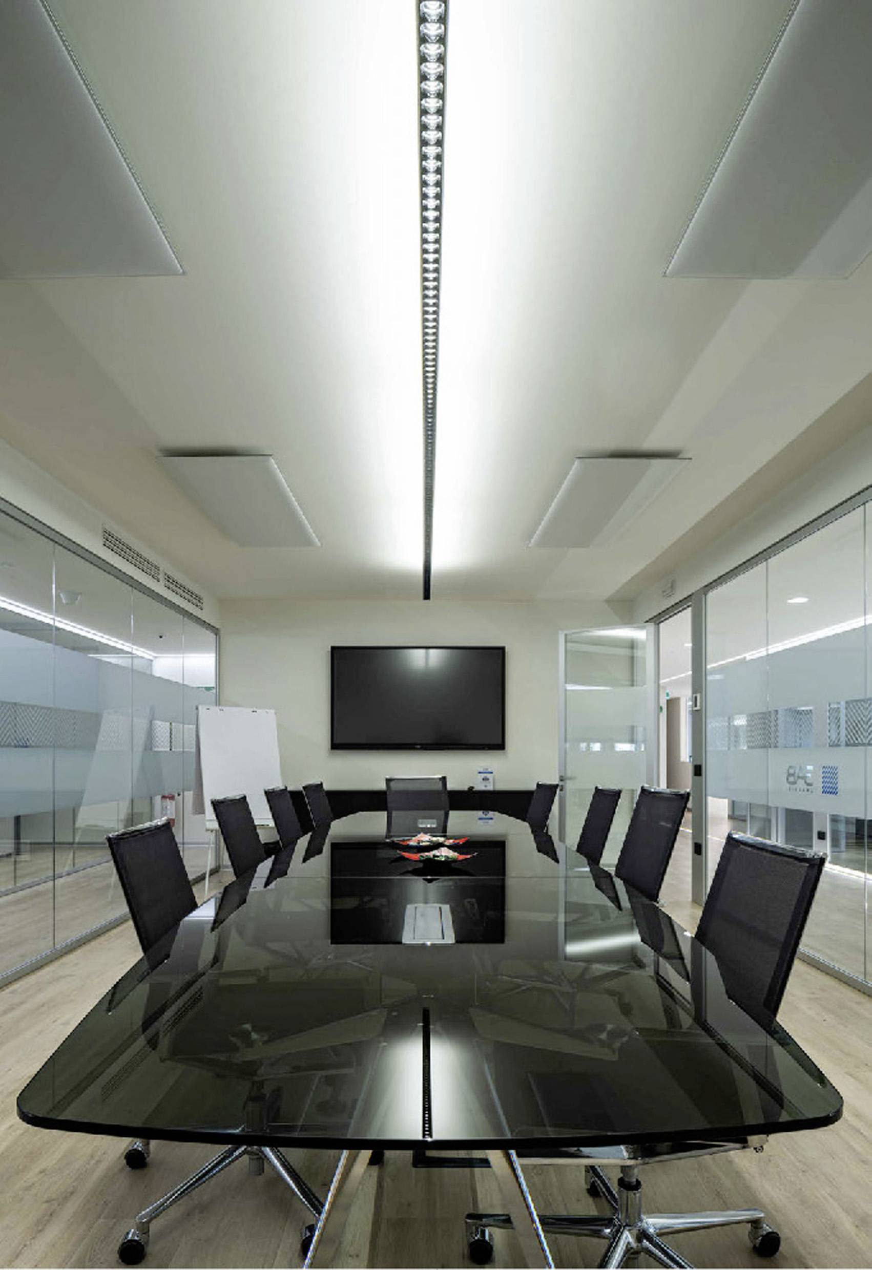 A modern meeting room with a long, black table, surrounded by several black chairs. A flat-screen display is mounted on the wall.