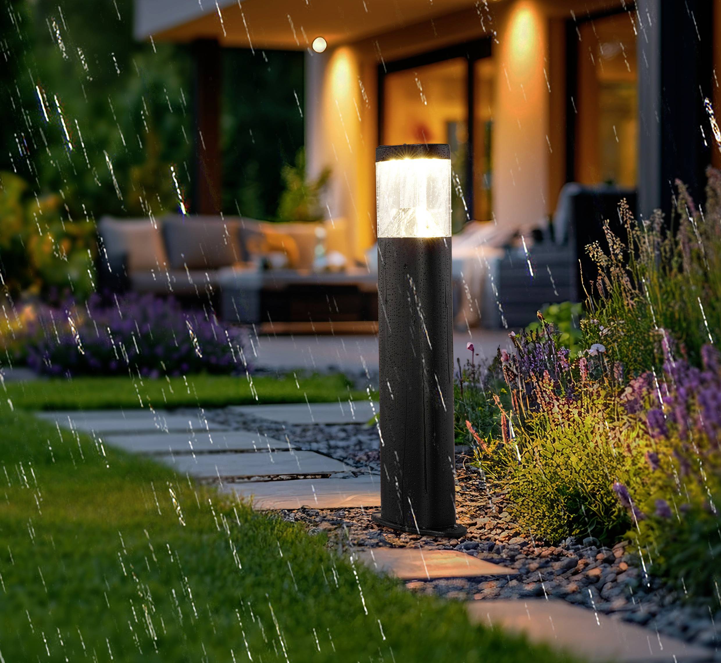 Outdoor modern garden with a glowing pathway light, surrounded by wet foliage and rain, leading to a warmly illuminated house.