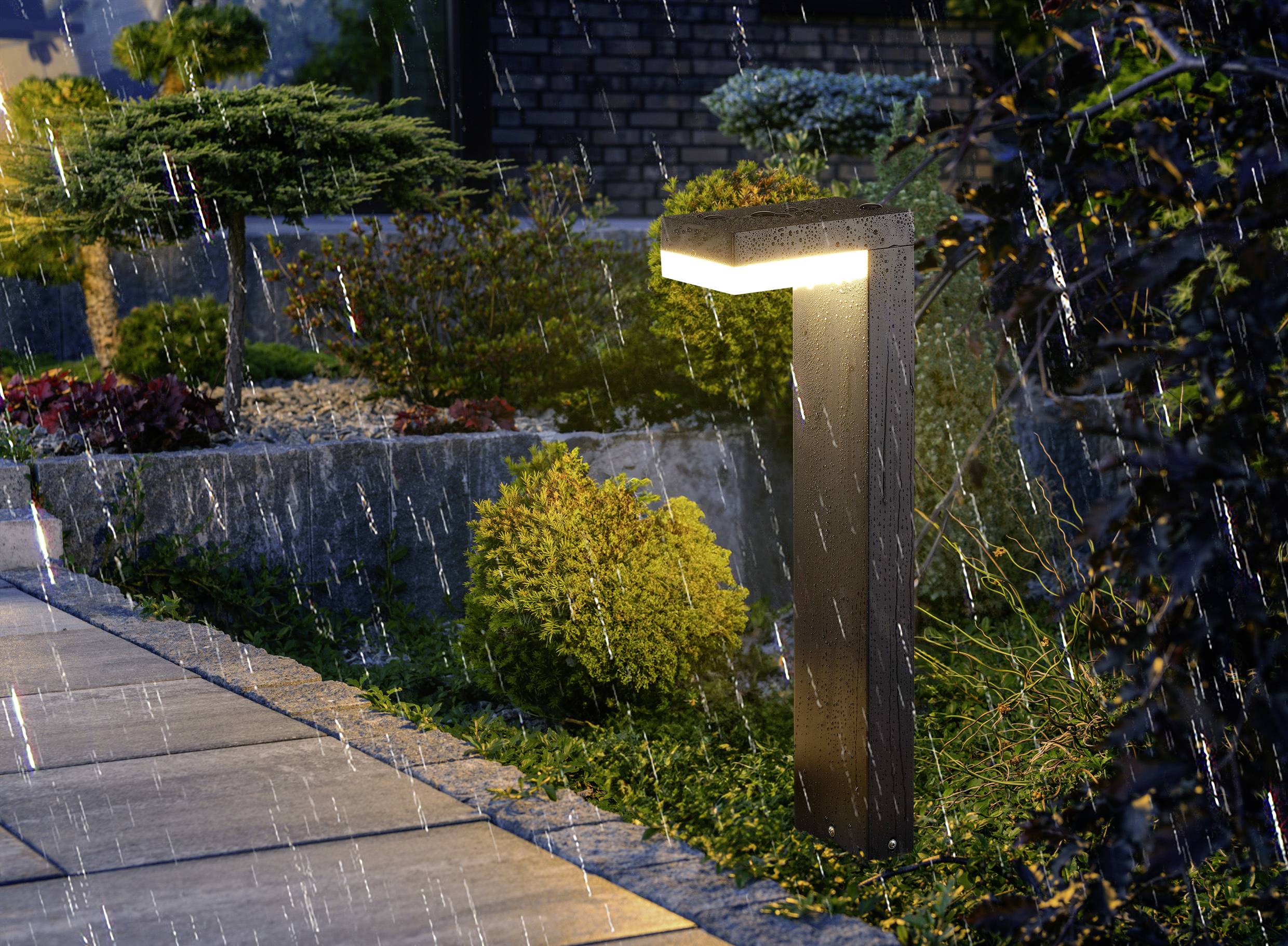 A modern garden light illuminates a rain-soaked pathway beside landscaped greenery, creating a serene night scene.
