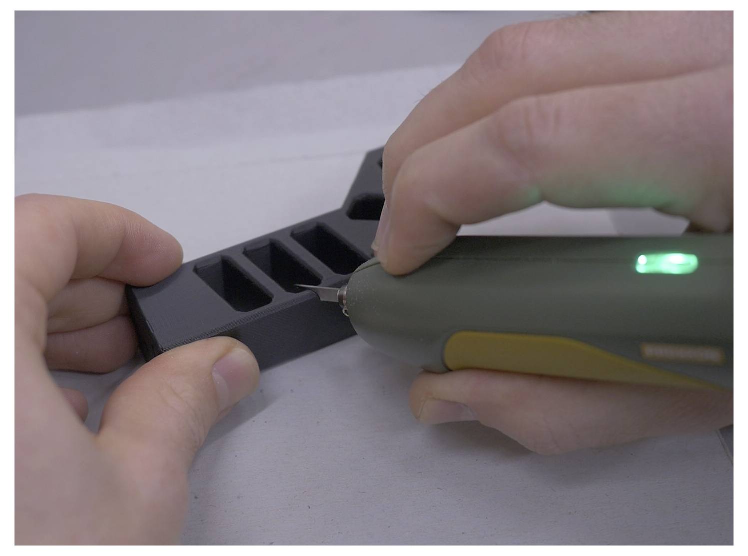 A hand holds a small electric tool, cutting into a rectangular piece of black material with multiple slots, on a light grey background.