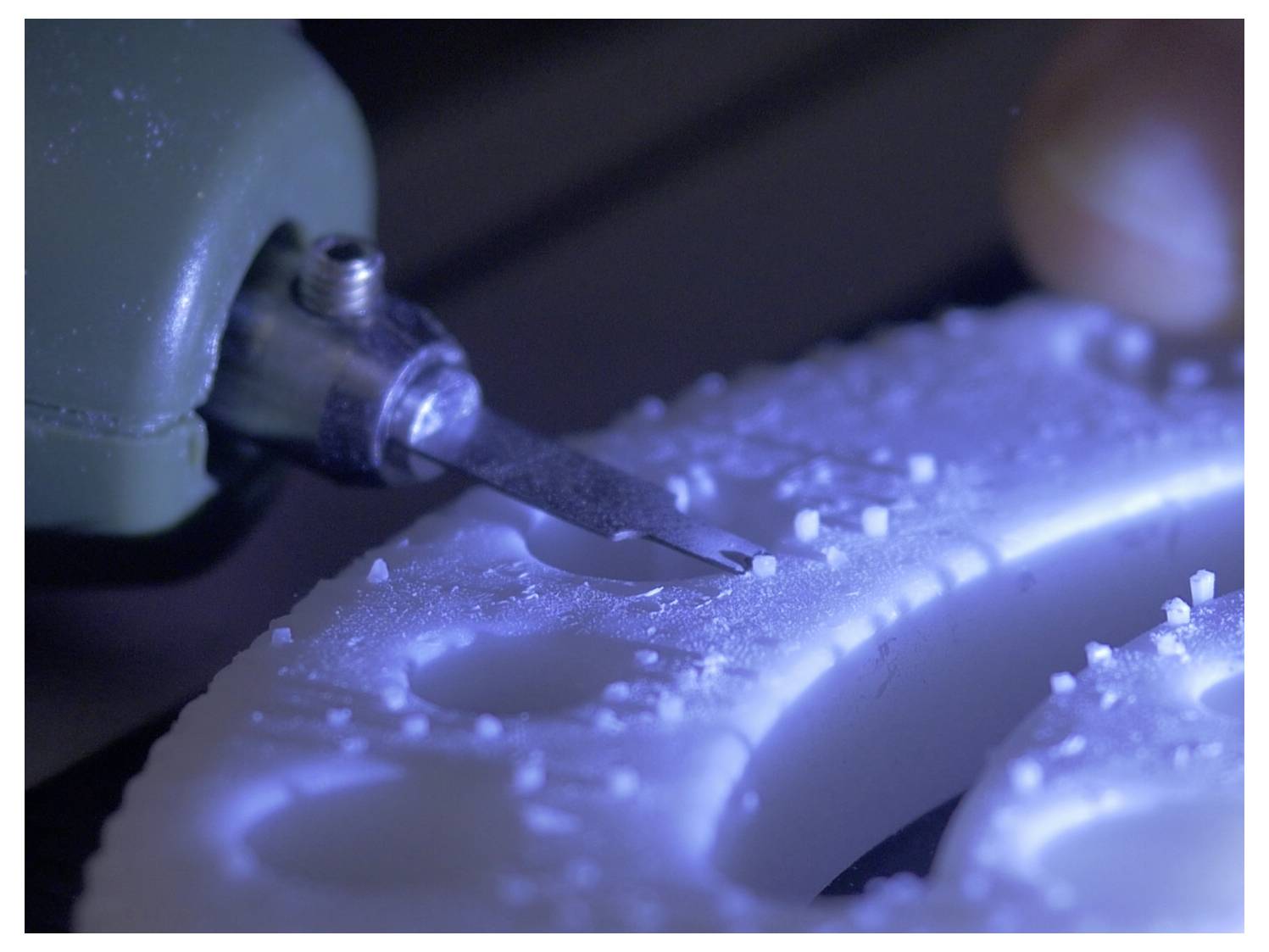 Close-up of a tool precisely cutting into a white material, possibly plastic, with scattered small debris around it, suggesting fine detailed work.