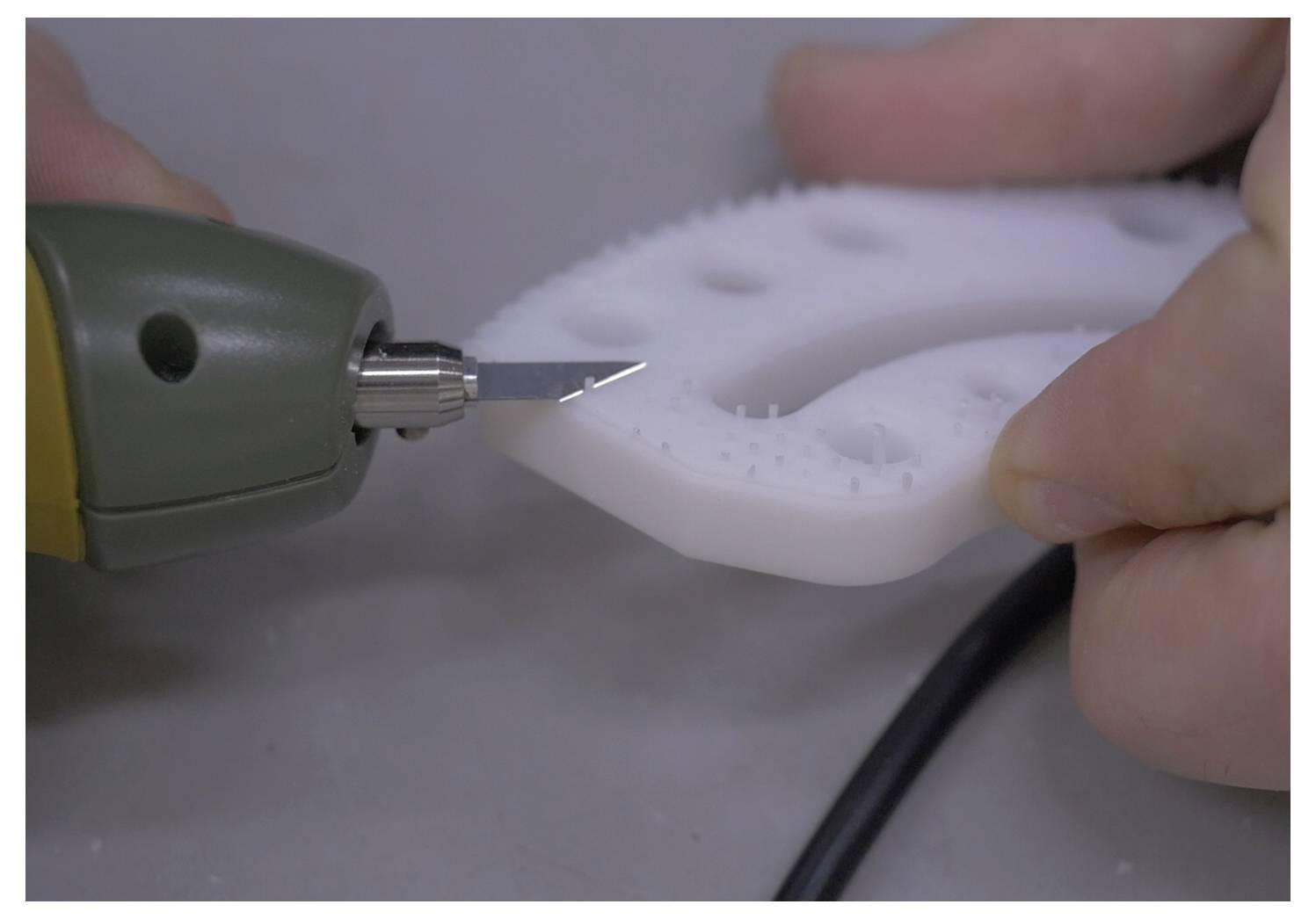 A close-up of a person using a heated wire cutter to cut a white foam sheet with holes. The sheet is held firmly by two fingers.