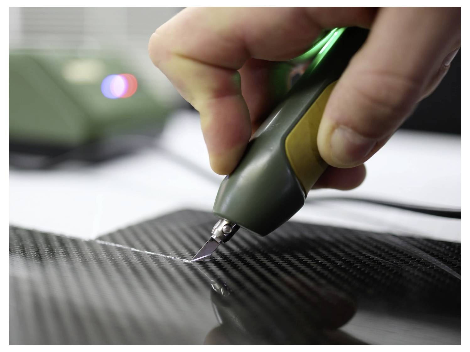 A hand holds a cutting tool, precisely carving into a sheet of carbon fiber material, showcasing precise craftsmanship and technology use.