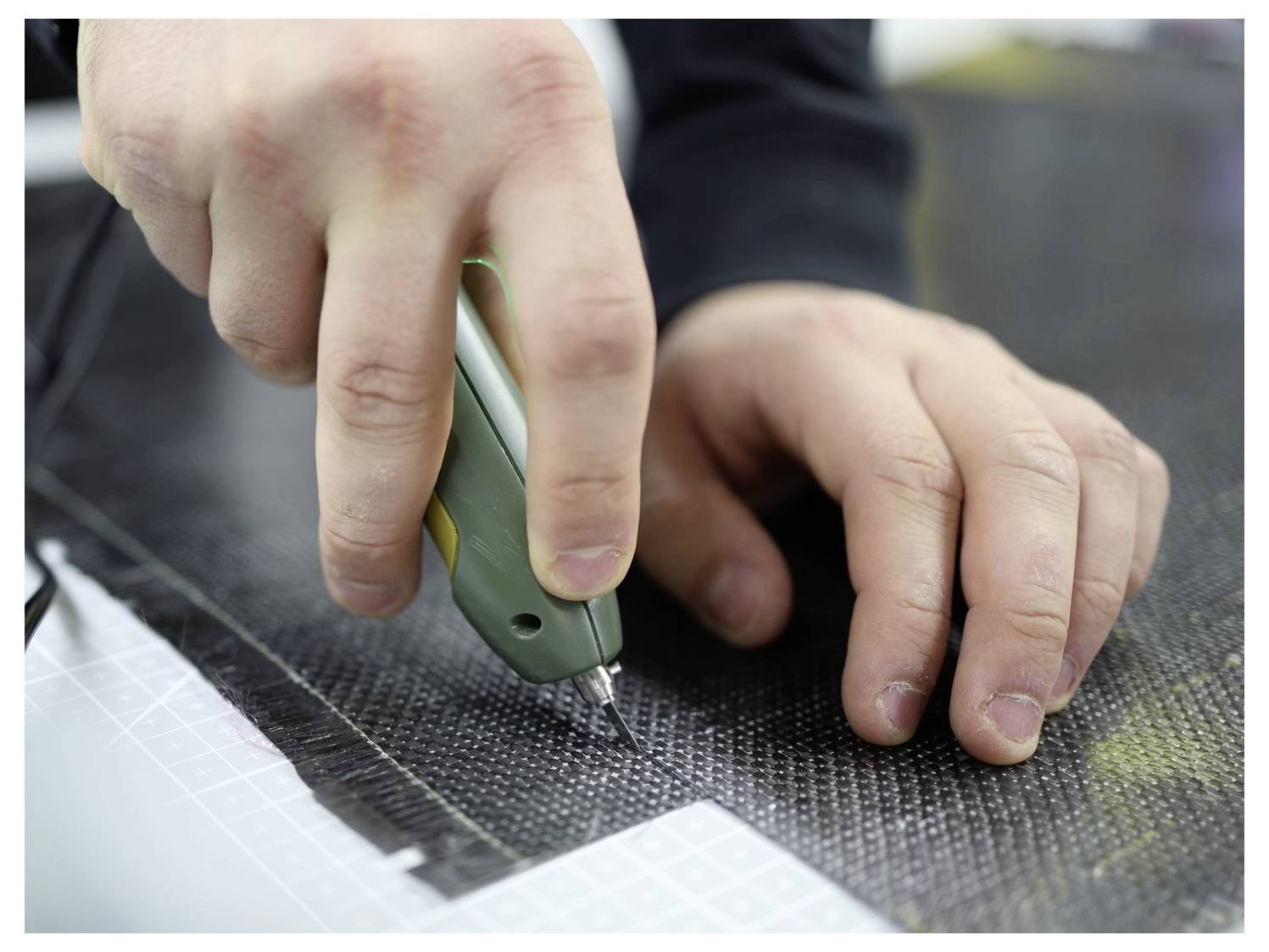 'Close-up of hands using a utility knife to cut a piece of black carbon fiber fabric on a cutting mat, focusing on precision work.'