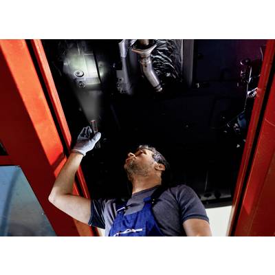A mechanic in blue overalls is inspecting the underside of a vehicle in a workshop, shining a torch.
