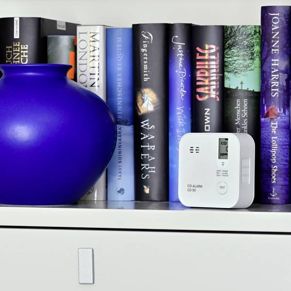 A carbon monoxide detector sits on a shelf next to a blue vase and books. Various authors' names can be read on the book spines.