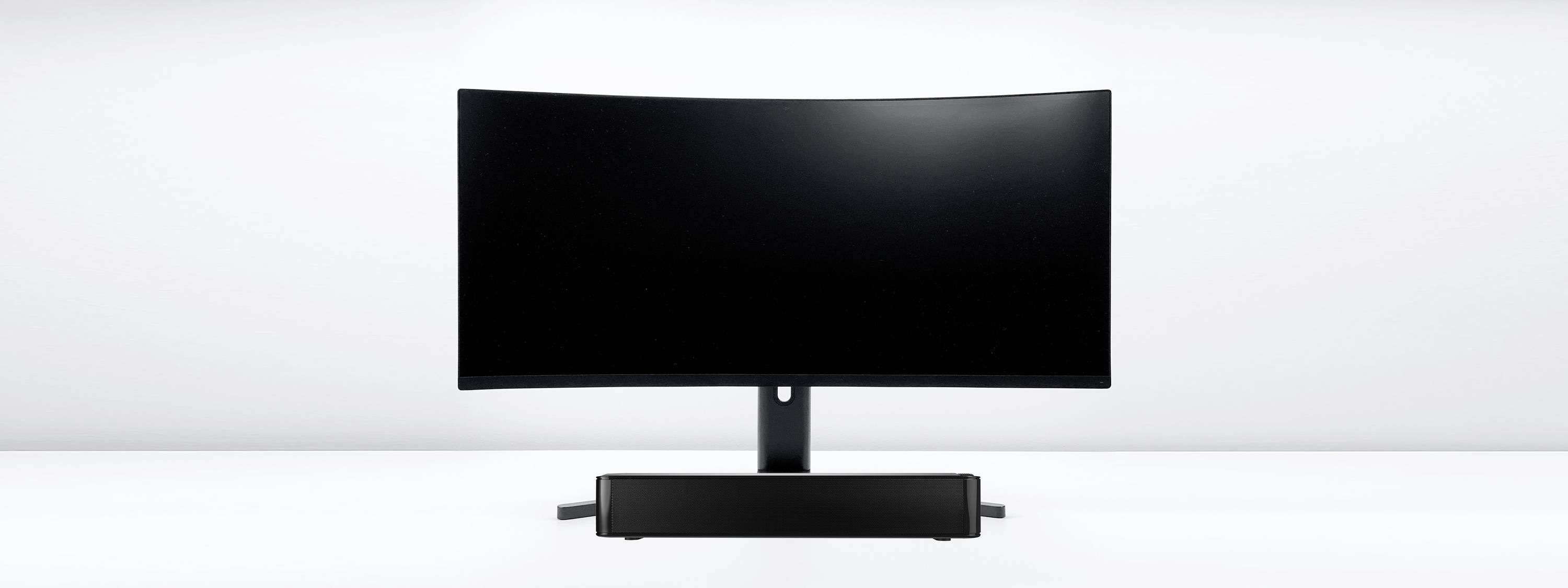 A curved, black computer monitor on a white table, with a black soundbar underneath, against a grey background.
