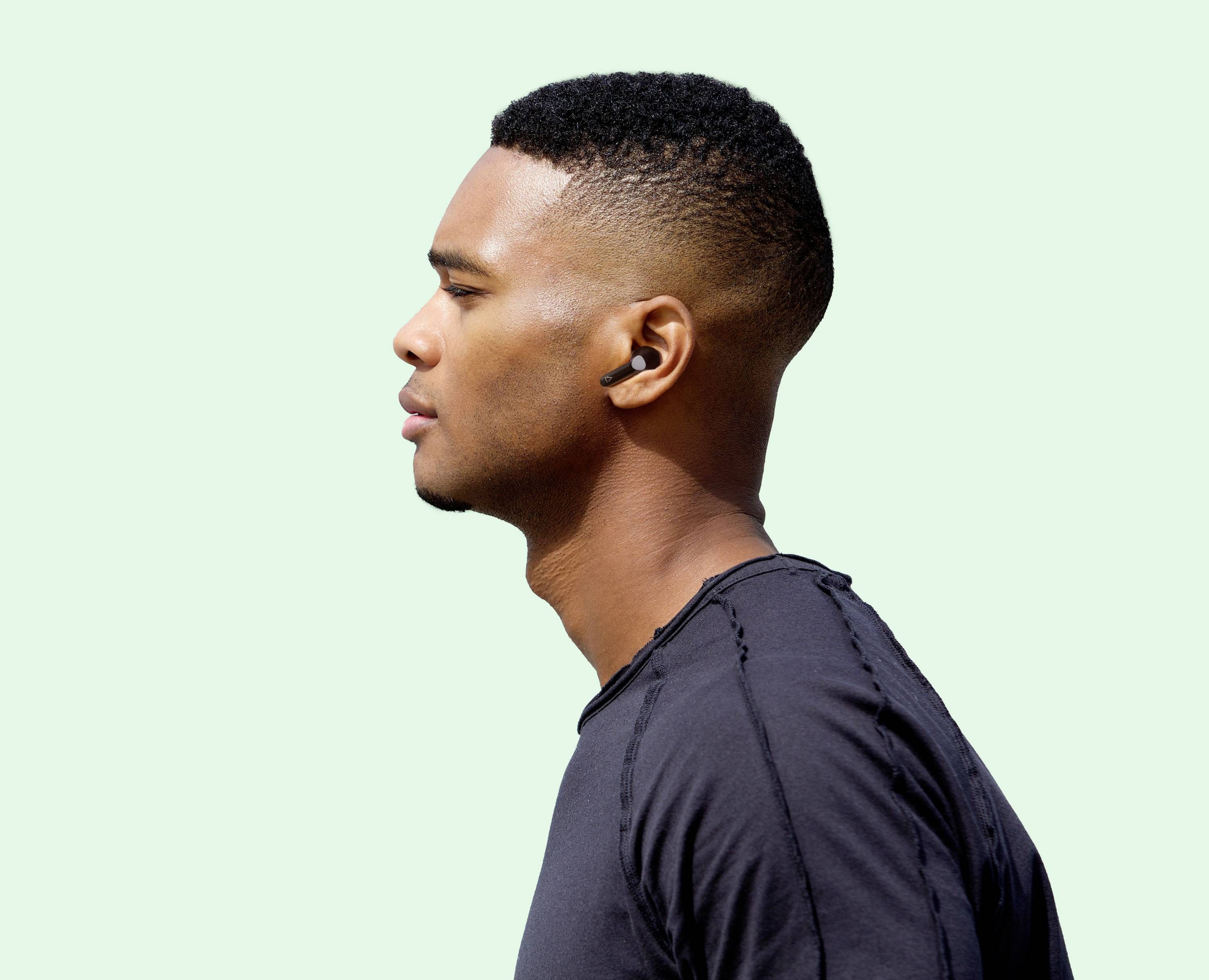 A person with short hair and earbuds is looking to the side, against a light green background. They are wearing a black t-shirt.