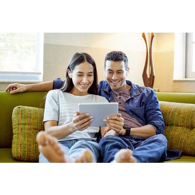 A couple sits relaxed on a sofa and smiles while looking at a tablet together. The background shows a lamp and windows.