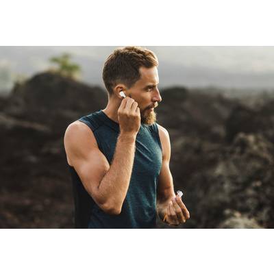 A man in a sleeveless sports vest is inserting wireless earbuds, standing outside in a rocky, natural setting.