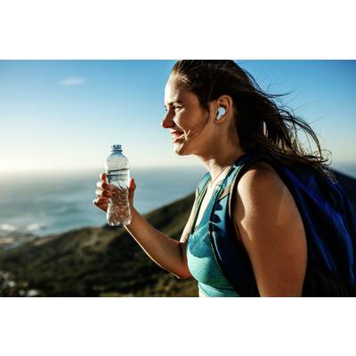 A woman hiking on a mountain path, holding a water bottle in her hand; sunny day, with nature in the background.