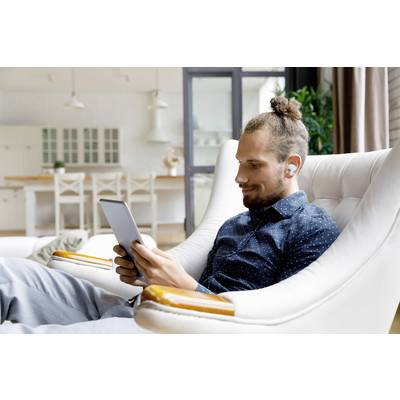 A man is sitting relaxed in an armchair and looking at a tablet. He is wearing wireless earbuds and is in a modernly furnished room.
