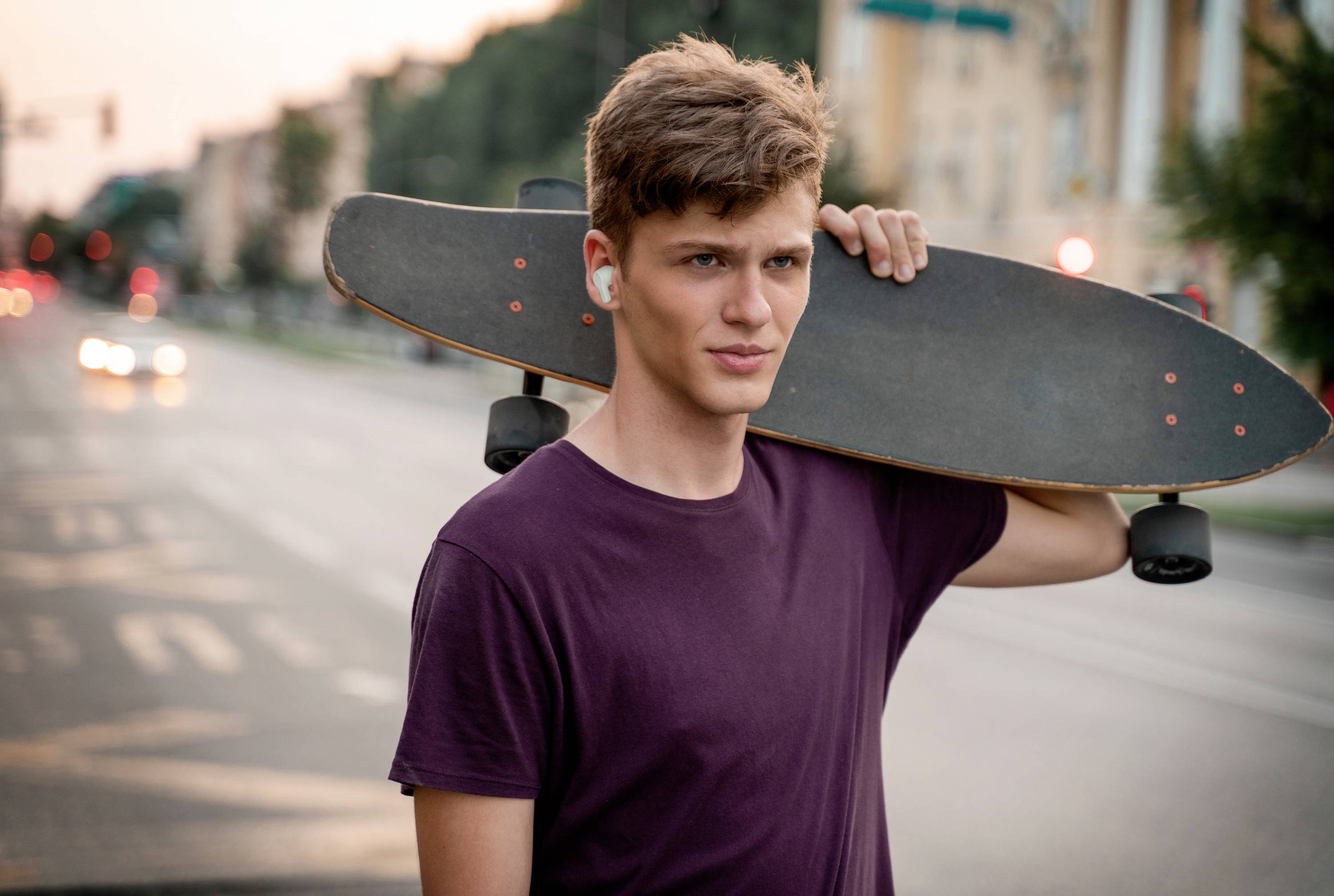 A young man with brown hair is carrying a skateboard on his shoulder and standing on a city street at sunset.