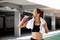 A woman in sportswear is drinking water from a bottle after her workout. She has a towel draped around her neck, in a car park.