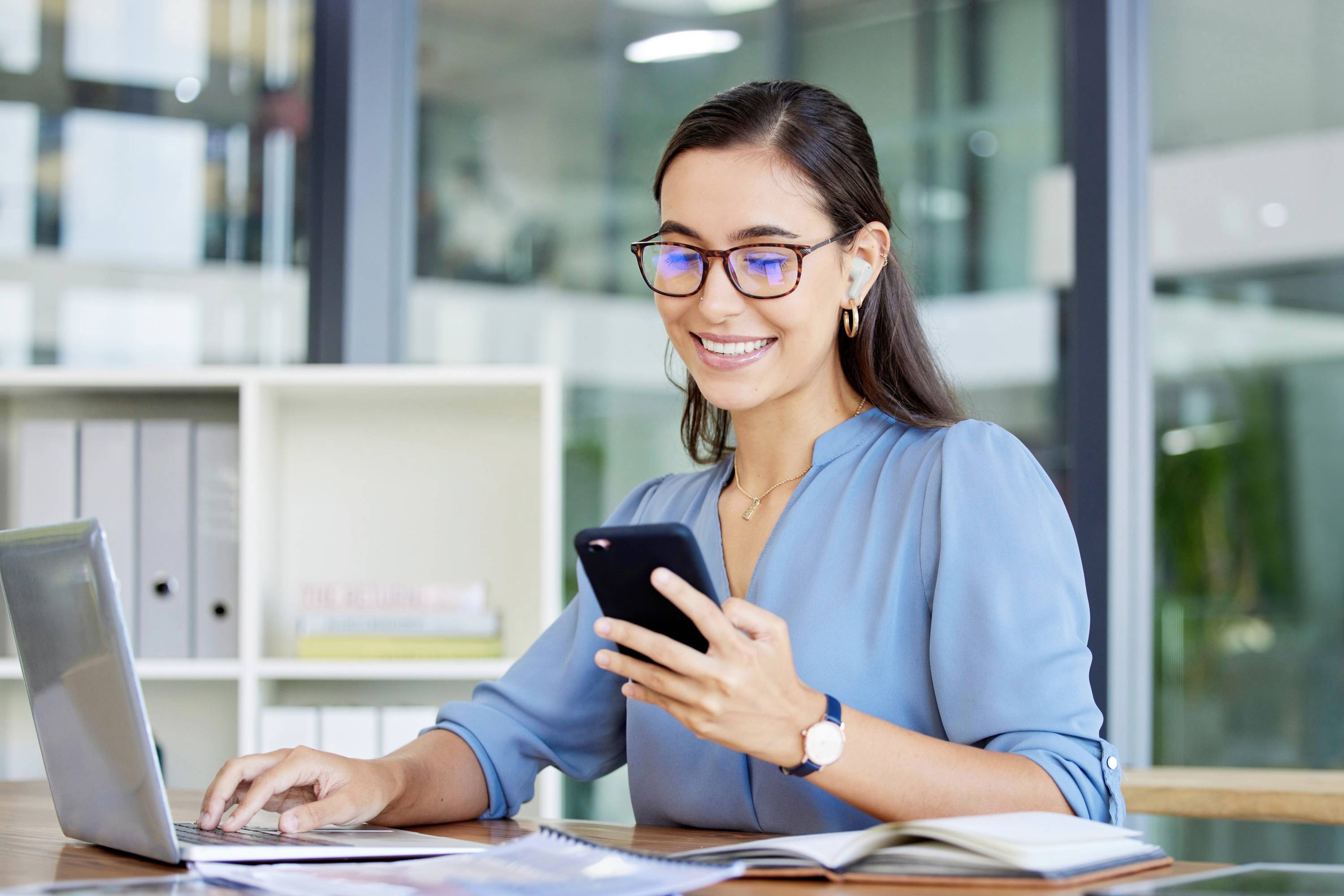 A smiling woman sits at a desk, looking at her mobile phone and typing on a laptop. An office can be seen in the background.