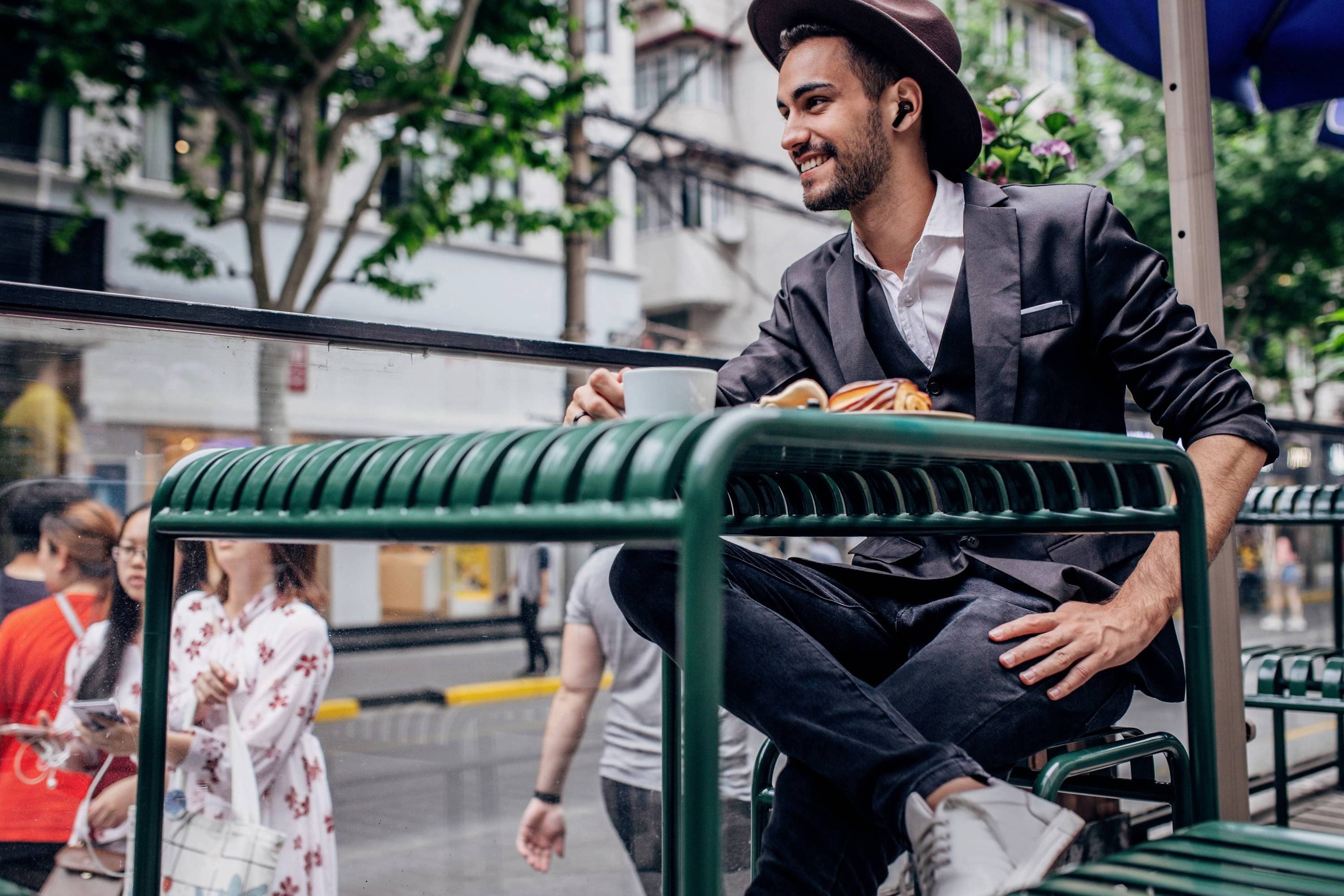 A man is sitting smiling in an outdoor café, drinking coffee. Urban street scenes with people can be seen in the background.