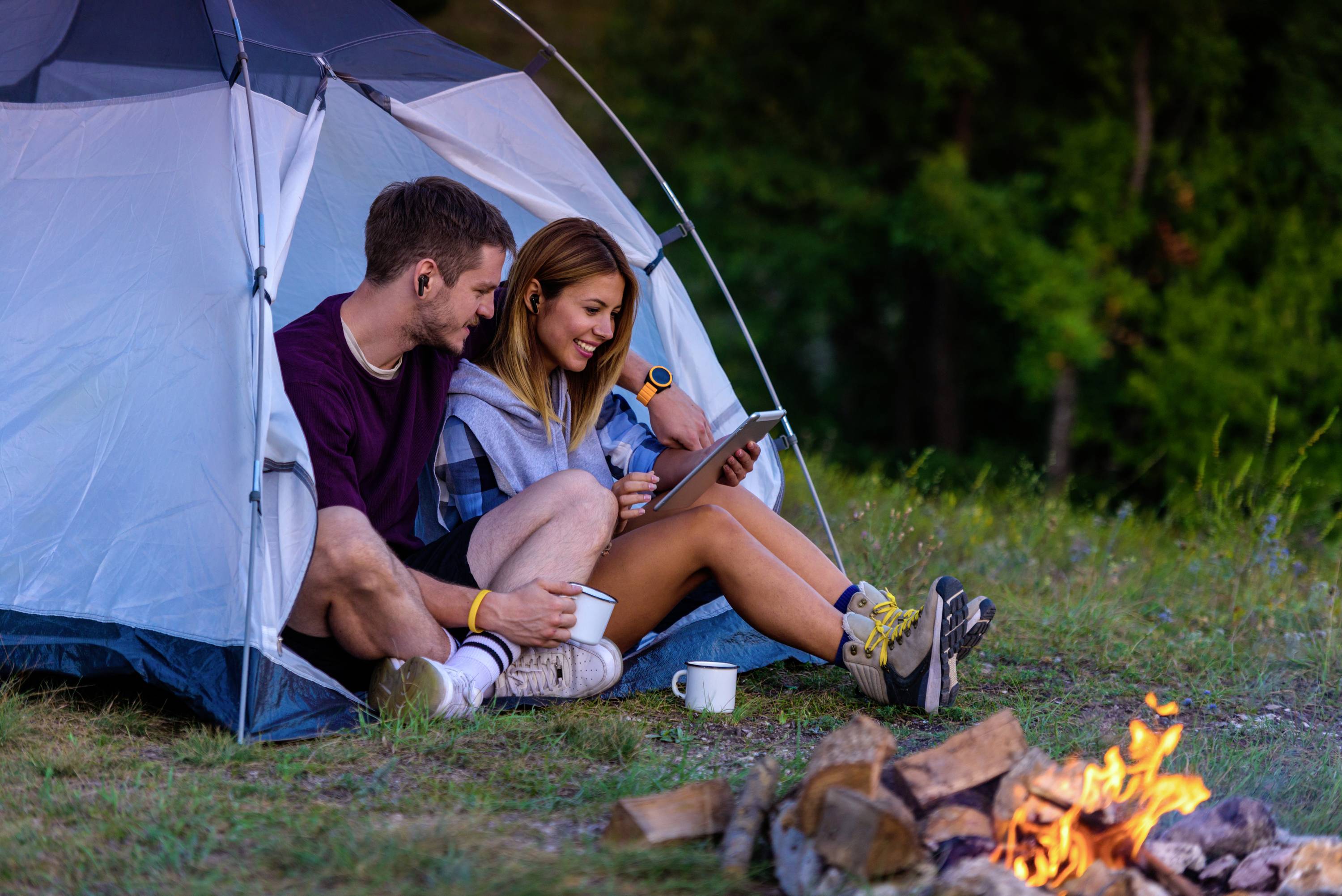 A couple are sitting in front of a tent, looking at a tablet whilst a campfire burns in the foreground. They are smiling and drinking coffee.