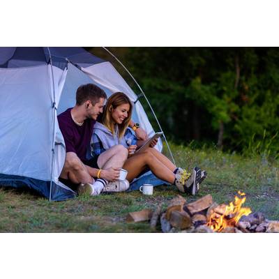 A couple are sitting in front of a tent, looking at a tablet whilst a campfire burns in the foreground. They are smiling and drinking coffee.