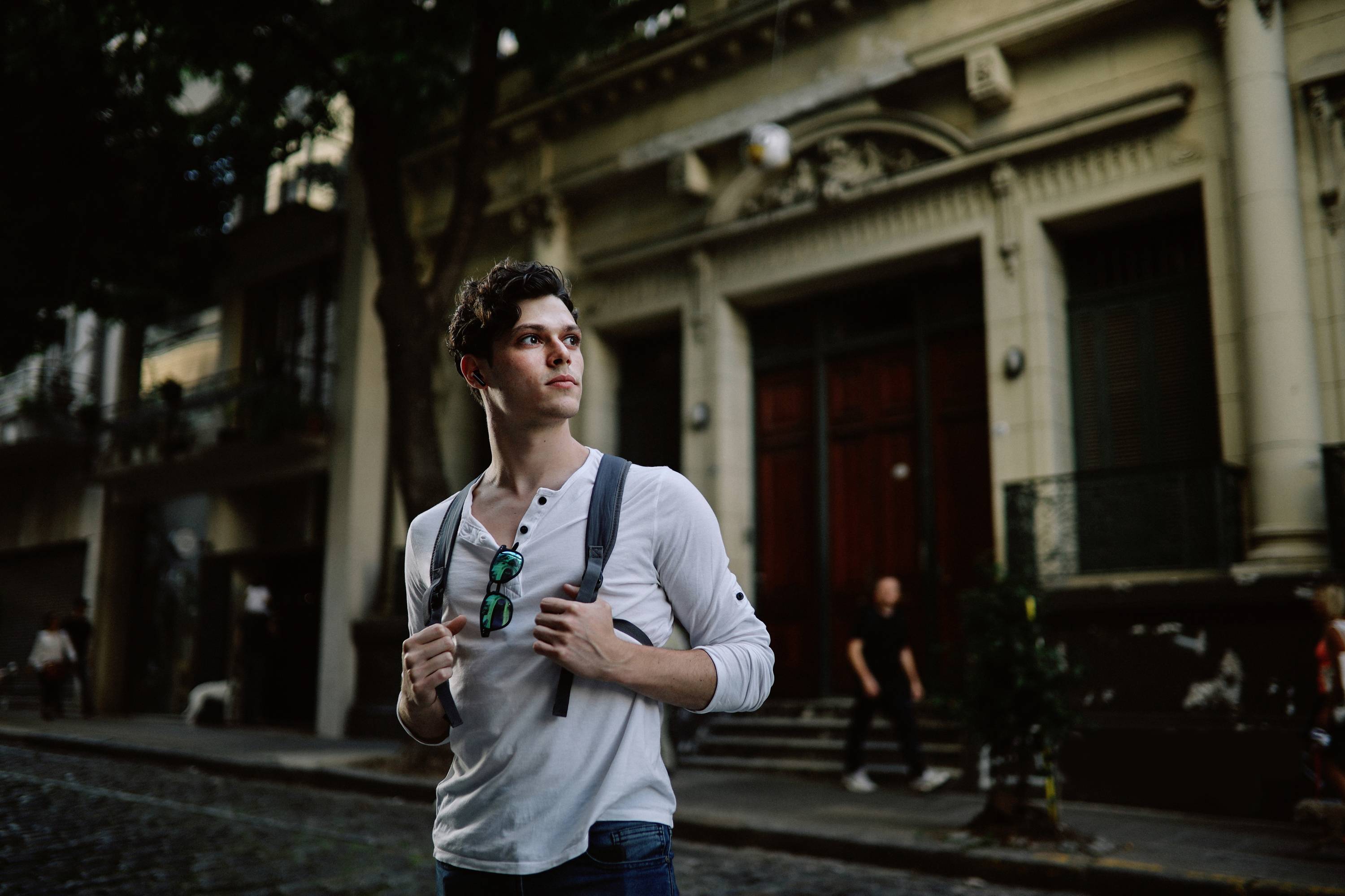 A man with a rucksack is standing on an urban street in front of an old building. He is looking to the side while people walk past in the background.