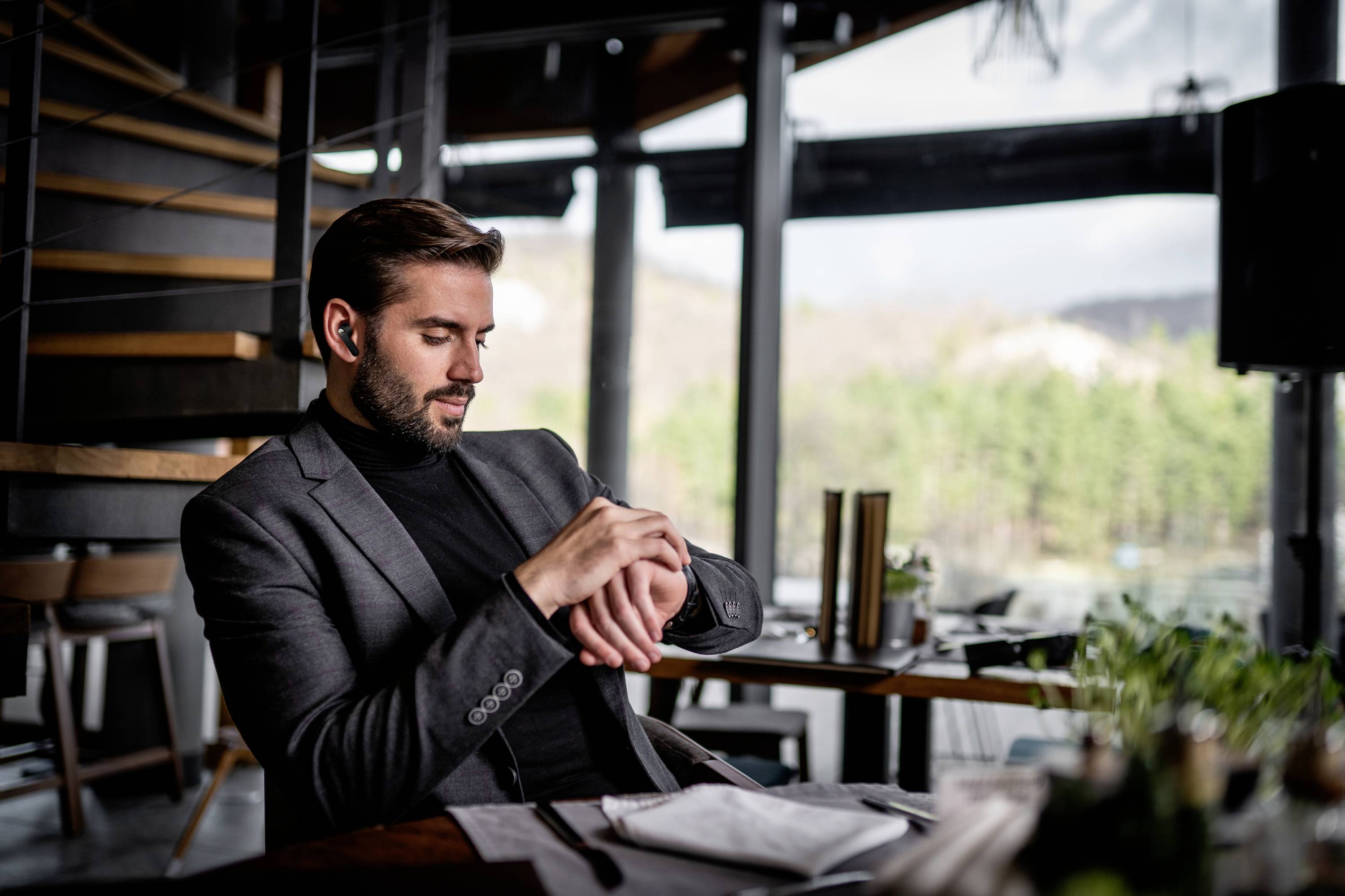 A man in a suit is sitting at a table in a modern restaurant, looking at his wristwatch. Nature can be seen in the background.