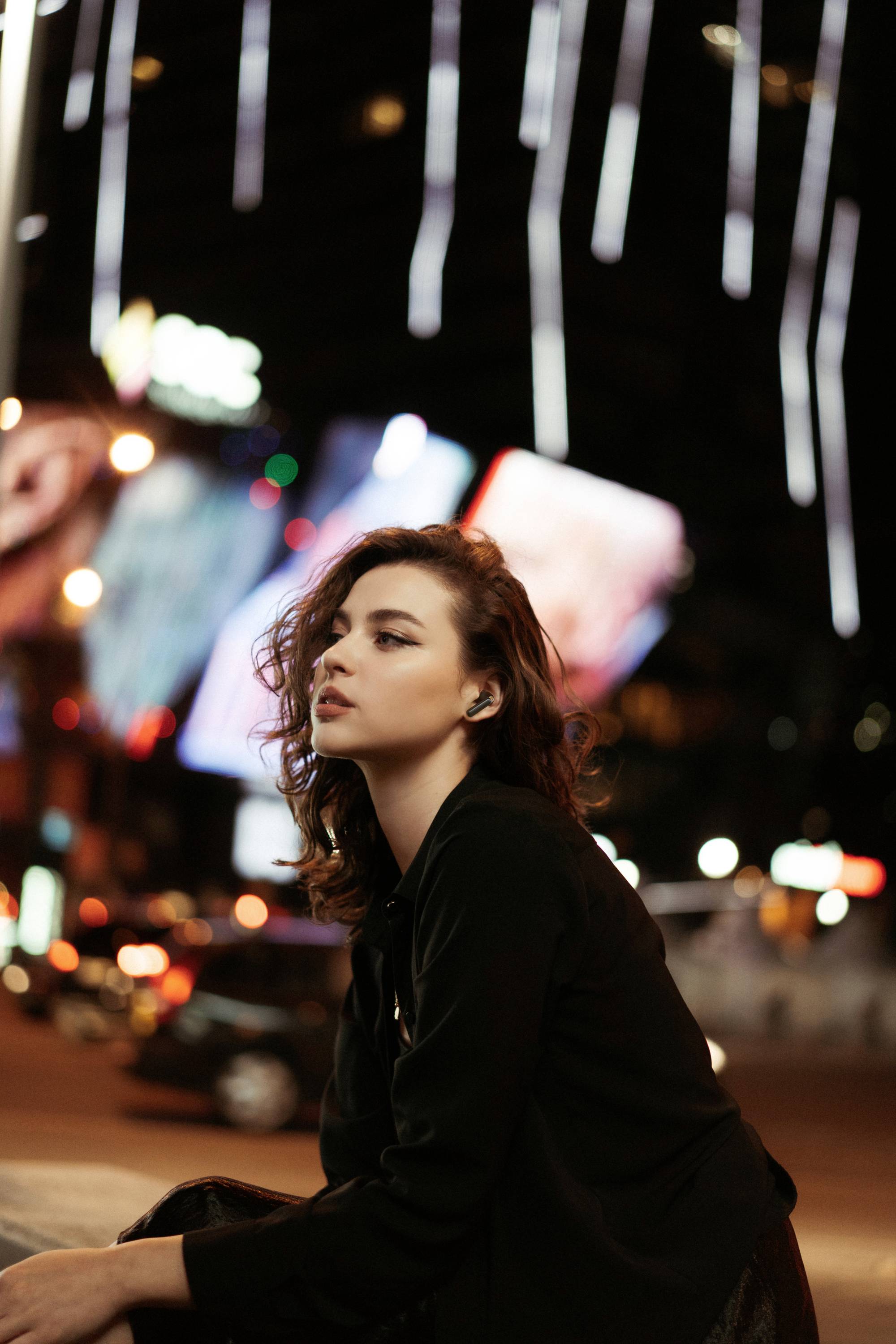A woman sits pensively on a street at night, surrounded by blurred lights and passing cars in the city.