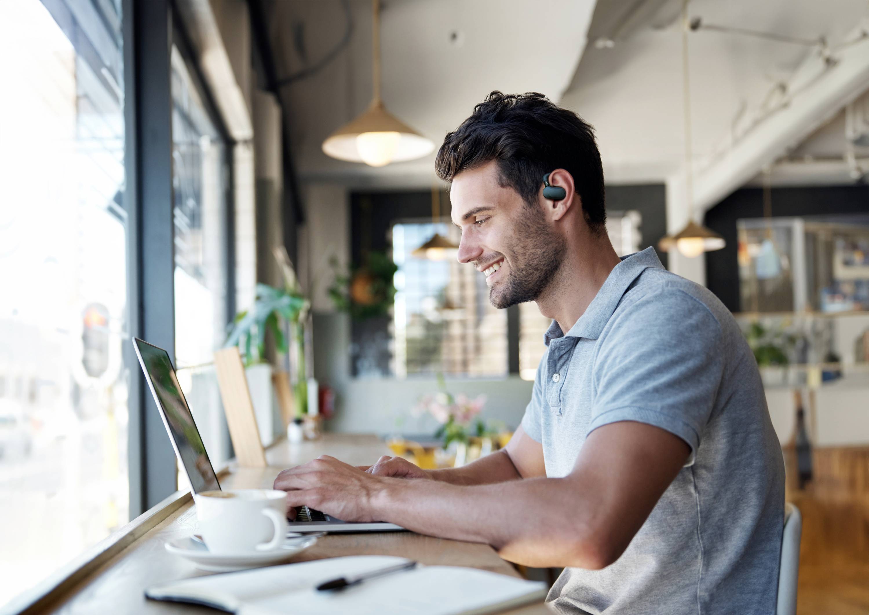 A man sits by a sunlight-filled café window, smiling as he works on a laptop and wearing wireless earbuds.