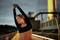 A woman in sportswear is stretching on a bridge at dusk. She is wearing wireless earbuds and appears focused and relaxed.