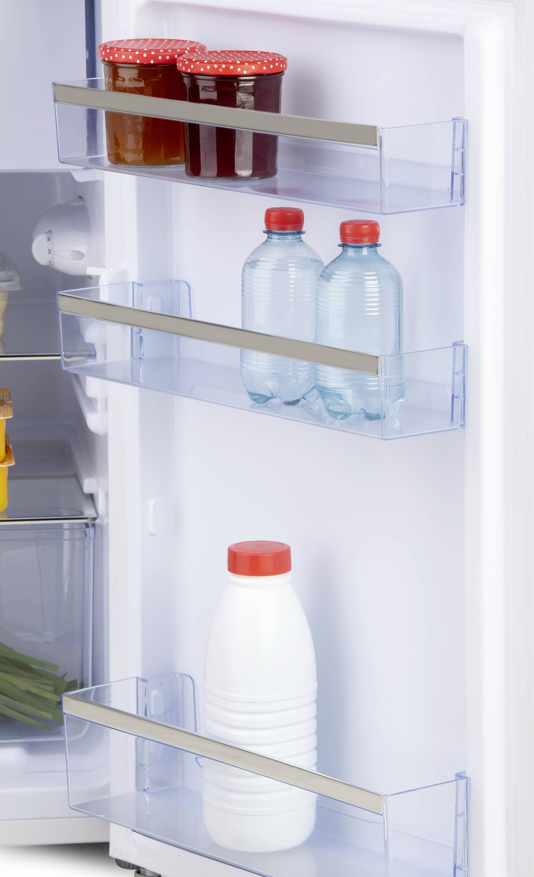 In an open fridge, there are shelves in the door with two bottles of water and a bottle of milk. On the top shelf, there are jars with red lids.