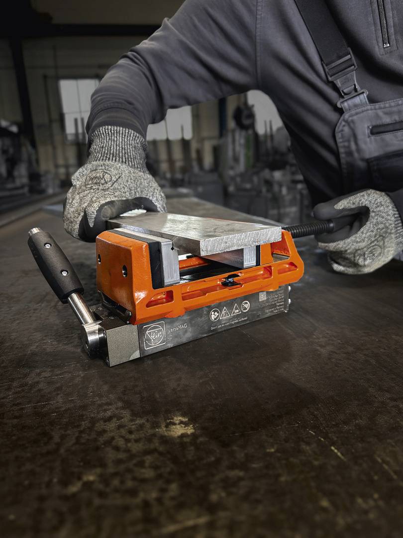 A person wearing protective gloves is holding an orange magnetic clamp tool that is securing a piece of metal on a table.