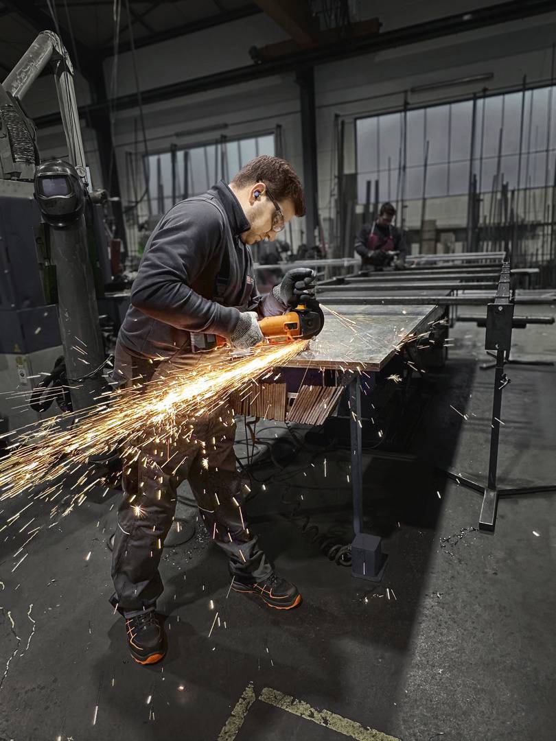 A person is grinding a metal piece with an angle grinder, sparks flying. In the background, a workshop hall.