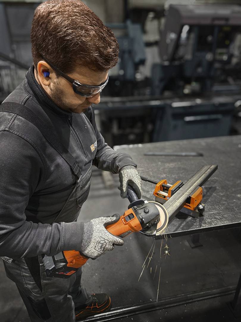 A man wearing safety glasses and ear plugs is grinding a metal pipe in a workshop. Sparks are flying from the grinding machine.