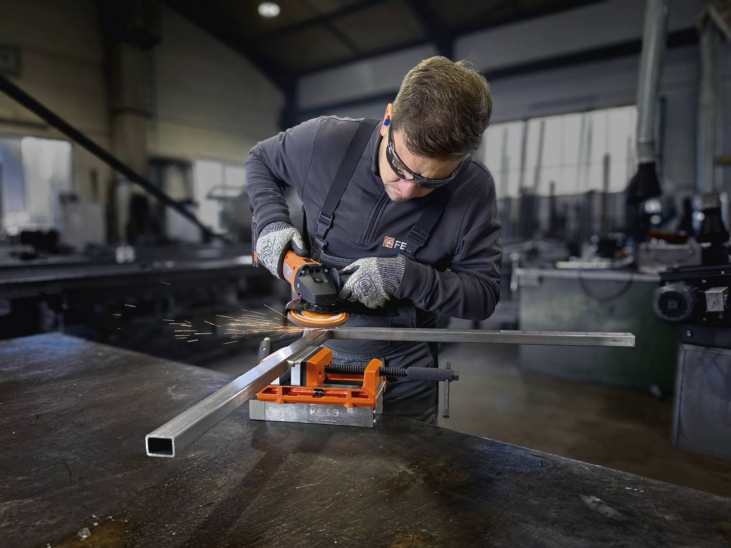 A person wearing safety goggles and gloves is grinding a metal rod with an angle grinder in a workshop. Sparks are flying.